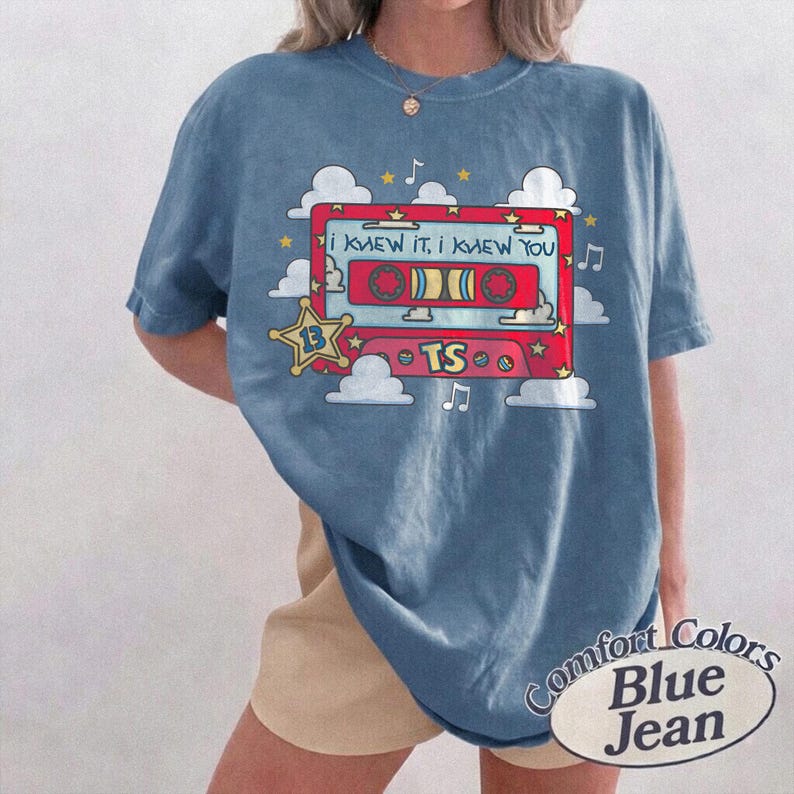 May include: A blue short-sleeve t-shirt featuring a graphic of a red cassette tape with the text "I KNEW IT, I KNEW YOU" and "TS". The design includes white clouds, yellow stars, and musical notes. The shirt has a crew neckline and a relaxed fit.