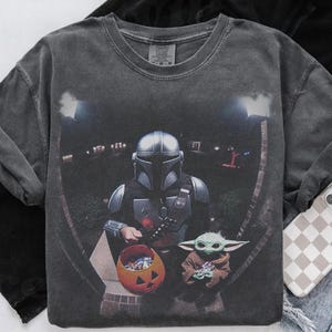 May include: Dark gray long-sleeve shirt featuring a graphic of The Mandalorian and Grogu trick-or-treating on Halloween. The Mandalorian is holding a pumpkin-shaped candy bucket. Grogu is holding a toy. The shirt has a vintage wash.