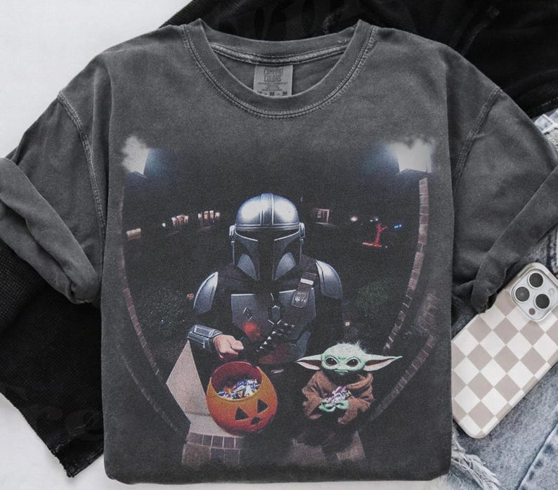 May include: Dark gray long-sleeve shirt featuring a graphic of The Mandalorian and Grogu trick-or-treating on Halloween. The Mandalorian is holding a pumpkin-shaped candy bucket. Grogu is holding a toy. The shirt has a vintage wash.