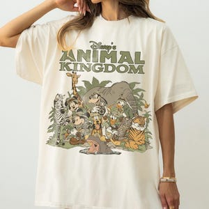 Puede incluir: Camiseta color crema con el texto "Disney's Animal Kingdom" en verde sobre un gr&aacute;fico de varios personajes y animales de Disney. El gr&aacute;fico incluye Mickey Mouse, Donald Duck y un tigre.
