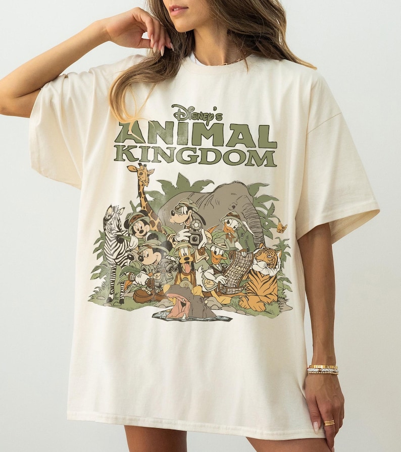 Puede incluir: Camiseta color crema con el texto "Disney's Animal Kingdom" en verde sobre un gr&aacute;fico de varios personajes y animales de Disney. El gr&aacute;fico incluye Mickey Mouse, Donald Duck y un tigre.