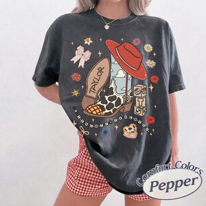 K&ouml;nnte beinhalten: Ein anthrazitfarbenes Kurzarm-T-Shirt mit einem Grafikdruck, der einen Cowboystiefel mit Kuhfleckenmuster und dem Namen TAYLOR, einen roten Cowboyhut, einen Stapel Bl&ouml;cke mit den Buchstaben T S, eine Schleife, Blumen und Sterne zeigt.