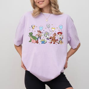 May include: A light purple t-shirt featuring a colorful graphic of animated characters holding balloons. The design includes a green dinosaur, a cowboy, a space ranger, a horse, and other characters. The shirt is a relaxed fit.