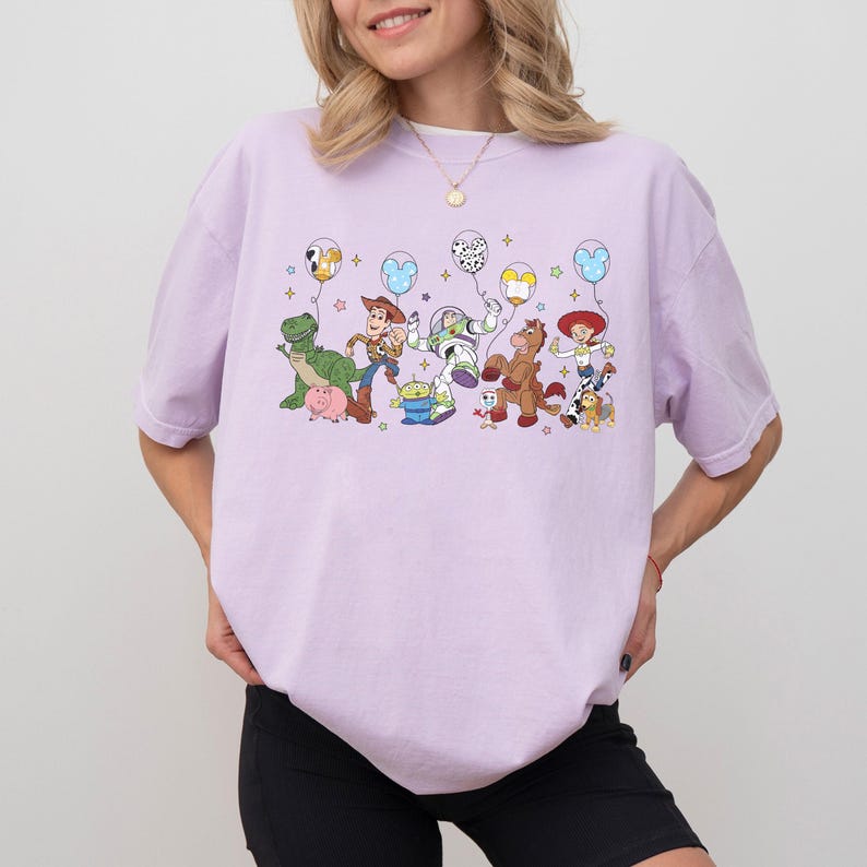May include: A light purple t-shirt featuring a colorful graphic of animated characters holding balloons. The design includes a green dinosaur, a cowboy, a space ranger, a horse, and other characters. The shirt is a relaxed fit.