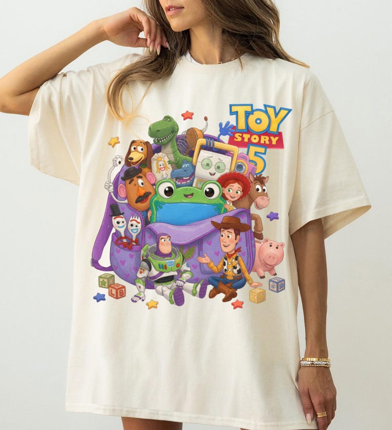 May include: An off-white t-shirt featuring a colorful graphic print of Toy Story 5 characters including Woody, Buzz Lightyear, and a green frog emerging from a purple backpack. The text Toy Story 5 is printed in blue and red above the characters.