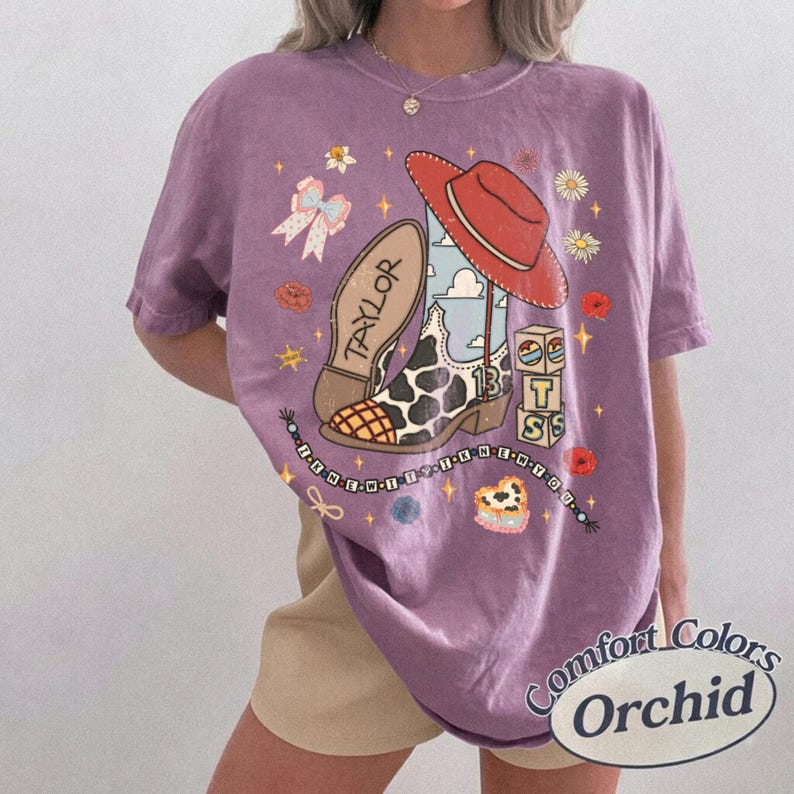 K&ouml;nnte beinhalten: Ein violettes Kurzarm-T-Shirt mit einem Grafikdruck, der einen Cowboystiefel mit Kuhmuster, einen roten Cowboyhut, eine Schleife, Blumen und W&uuml;rfel mit den Buchstaben T, S und 13 zeigt. Der Schriftzug TAYLOR befindet sich auf dem Stiefel.