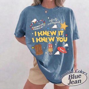 May include: A blue t-shirt featuring a graphic print with the text "I KNEW IT I KNEW YOU", "infinity and beyond", and "YOU'VE GOT A FRIEND IN ME". The design includes illustrations of cowboy boots, a red hat, alphabet blocks, stars, and clouds.