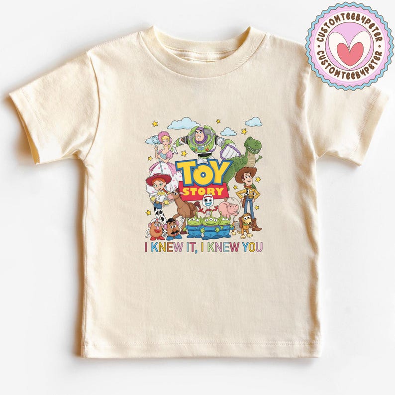 May include: A cream-colored short-sleeve t-shirt featuring a colorful graphic of Toy Story characters including Woody, Buzz Lightyear, Jessie, and others. The text "TOY STORY" is printed in the center, with "I KNEW IT, I KNEW YOU" below the characters.