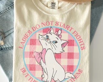 May include: Cream-colored t-shirt featuring a cartoon cat with a pink bow and the text "LADIES DO NOT START FIGHTS BUT THEY CAN FINISH THEM!". The design is set against a pink and white checkered background.