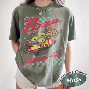 Pu&ograve; includere: T-shirt verde oliva con una grafica raffigurante tre alieni dei cartoni animati su un razzo, una pizza e la scritta "Pizza H-O-T-O". La maglietta ha anche la scritta "You can take me H-O-T-O Go".
