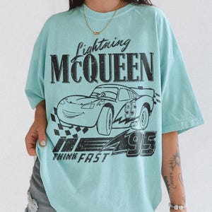 May include: A pale blue t-shirt with a black graphic of Lightning McQueen, the race car from the film Cars. The design includes the text "Lightning McQueen", the number "95", and "Think Fast".