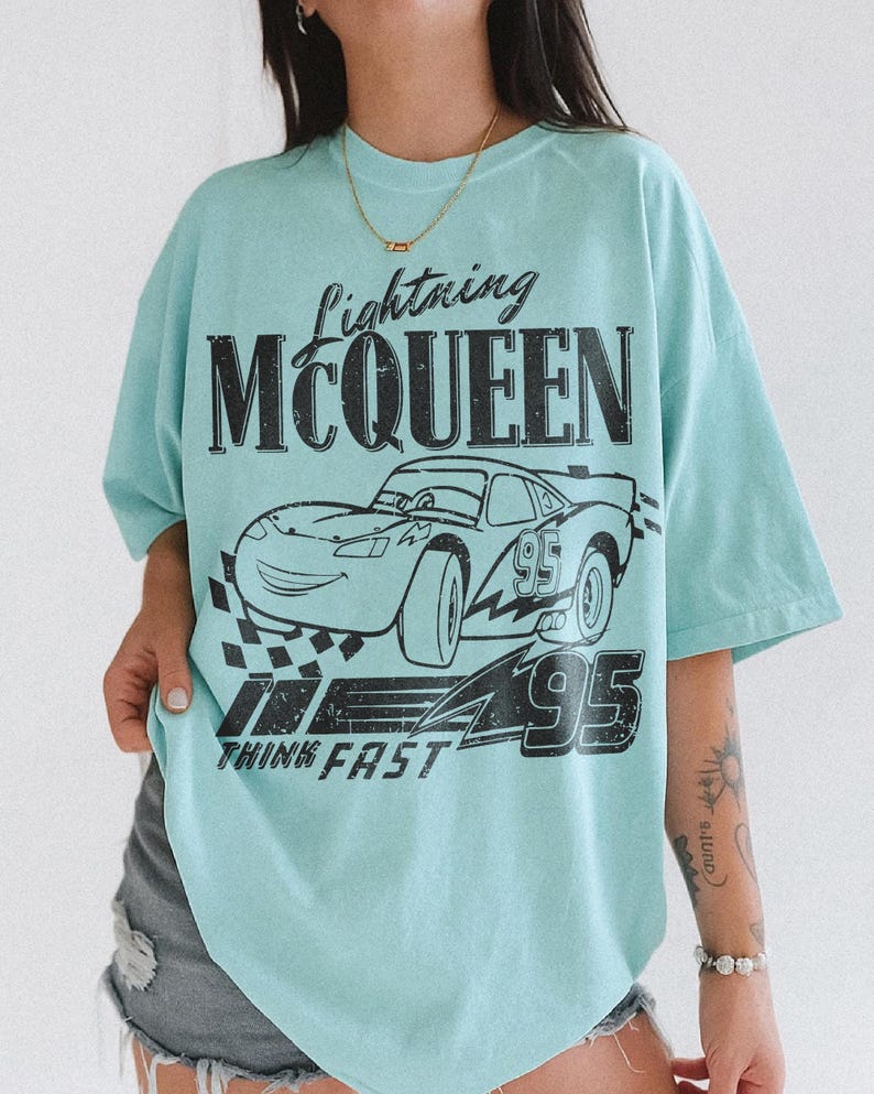 May include: A pale blue t-shirt with a black graphic of Lightning McQueen, the race car from the film Cars. The design includes the text "Lightning McQueen", the number "95", and "Think Fast".
