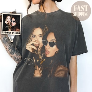 Puede incluir: Una camiseta gris carbón de cuello redondo con un gran estampado gráfico de dos personas sosteniendo copas de vino. Una persona tiene el pelo largo y bebe, mientras que la otra lleva gafas de sol oscuras. El estampado tiene una estética vintage.