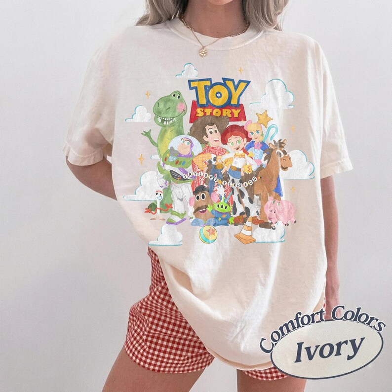 May include: An ivory short-sleeve t-shirt featuring a colorful graphic print of Toy Story characters including Woody, Buzz Lightyear, Rex, and Jessie. The design includes the Toy Story logo, clouds, and stars. The shirt is paired with red and white gingham shorts.