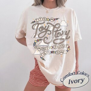 May include: An ivory short-sleeve t-shirt featuring a graphic print with the text "ITS A Toy Story BABY JUST YES SAY". The design includes rope-style lettering, colorful beads, small clouds, stars, and musical notes. The shirt has a crew neckline.