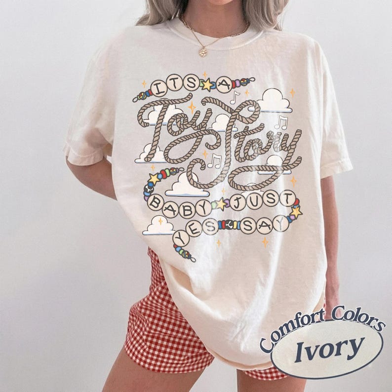 May include: An ivory short-sleeve t-shirt featuring a graphic print with the text "ITS A Toy Story BABY JUST YES SAY". The design includes rope-style lettering, colorful beads, small clouds, stars, and musical notes. The shirt has a crew neckline.