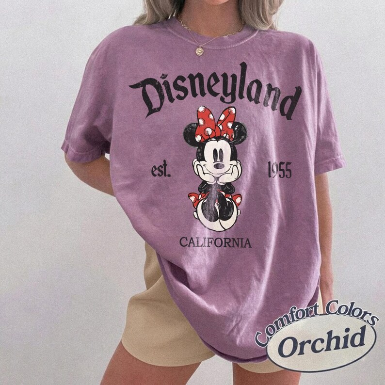 Może przedstawiać: Fioletowa koszulka z nadrukiem Myszki Minnie z czerwoną kokardą i napisem "Disneyland California est. 1955." Koszulka marki Comfort Colors.