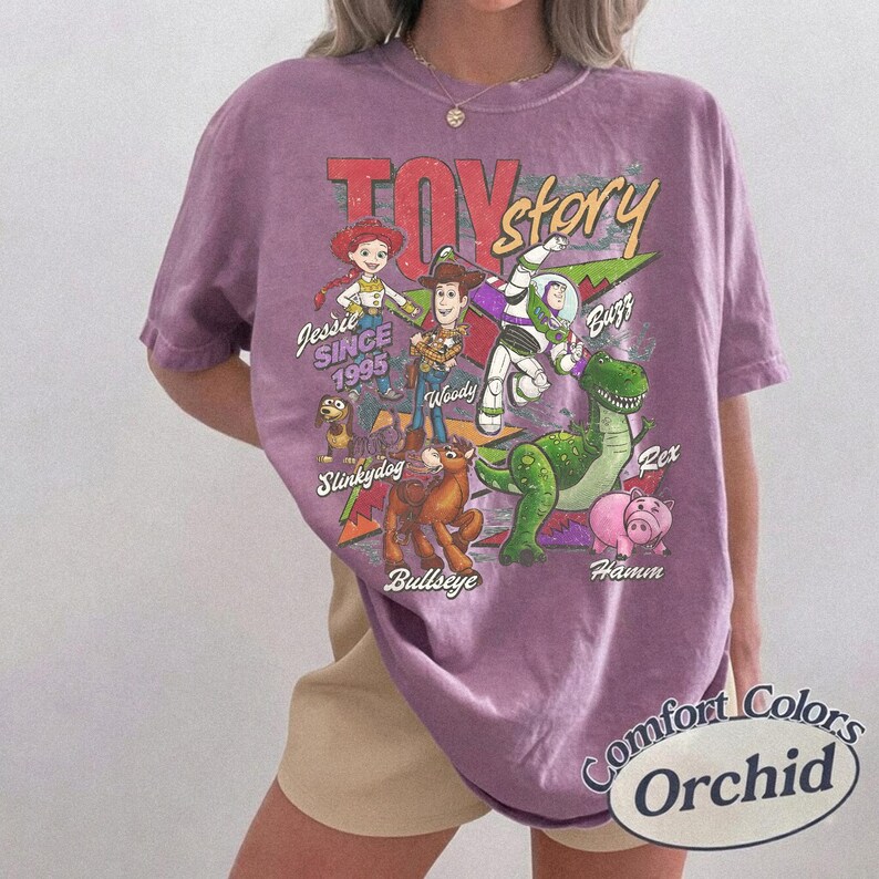 May include: A purple short-sleeve t-shirt featuring a graphic print of Toy Story characters including Jessie, Woody, Buzz Lightyear, Slinky Dog, Bullseye, Rex, and Hamm. The text reads TOY story, SINCE 1995, and the names of each character.