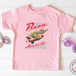 Pu&ograve; includere: Maglietta rosa chiaro con una grafica di tre alieni verdi in una navicella spaziale, le parole "Pizza Hot to Go" e una pizza su un pianeta. &Egrave; incluso anche il testo "You can take me".