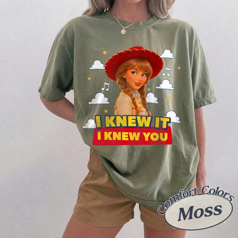 May include: A moss green short-sleeve t-shirt featuring a graphic of a character with braided hair wearing a red cowboy hat. The design includes white clouds, yellow stars, musical notes, and the text I KNEW IT I KNEW YOU in yellow and red.
