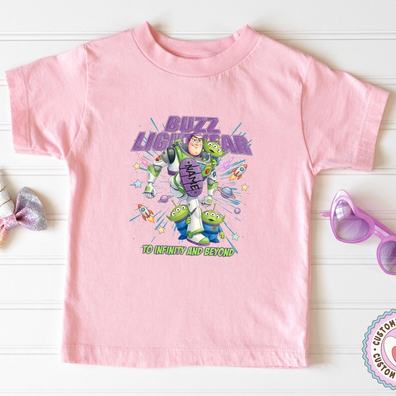 May include: A light pink short-sleeve t-shirt featuring a graphic of Buzz Lightyear and three green aliens from Toy Story. The design includes the text BUZZ LIGHTYEAR, a purple oval with the word NAME, and the phrase TO INFINITY AND BEYOND.