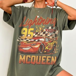 May include: Olive green t-shirt featuring a graphic of Lightning McQueen from the movie Cars. The design includes the text "Lightning 95" and "McQueen" in a vintage style. The shirt has short sleeves and a relaxed fit.