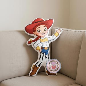 Peut inclure: Un coussin en forme du personnage Jessie de Toy Story. Le coussin repr&eacute;sente Jessie avec son chapeau rouge, ses cheveux roux tress&eacute;s, sa chemise blanche, son jean bleu et ses chaps &agrave; imprim&eacute; vache. Un autocollant circulaire avec du texte est visible.