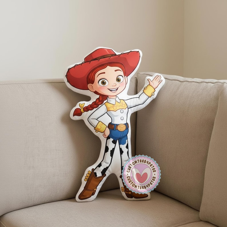 Peut inclure: Un coussin en forme du personnage Jessie de Toy Story. Le coussin repr&eacute;sente Jessie avec son chapeau rouge, ses cheveux roux tress&eacute;s, sa chemise blanche, son jean bleu et ses chaps &agrave; imprim&eacute; vache. Un autocollant circulaire avec du texte est visible.