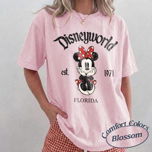 May include: A light pink t-shirt featuring a Minnie Mouse graphic with a red bow and the text "Disneyworld est. 1971 Florida". The shirt is from Comfort Colors.