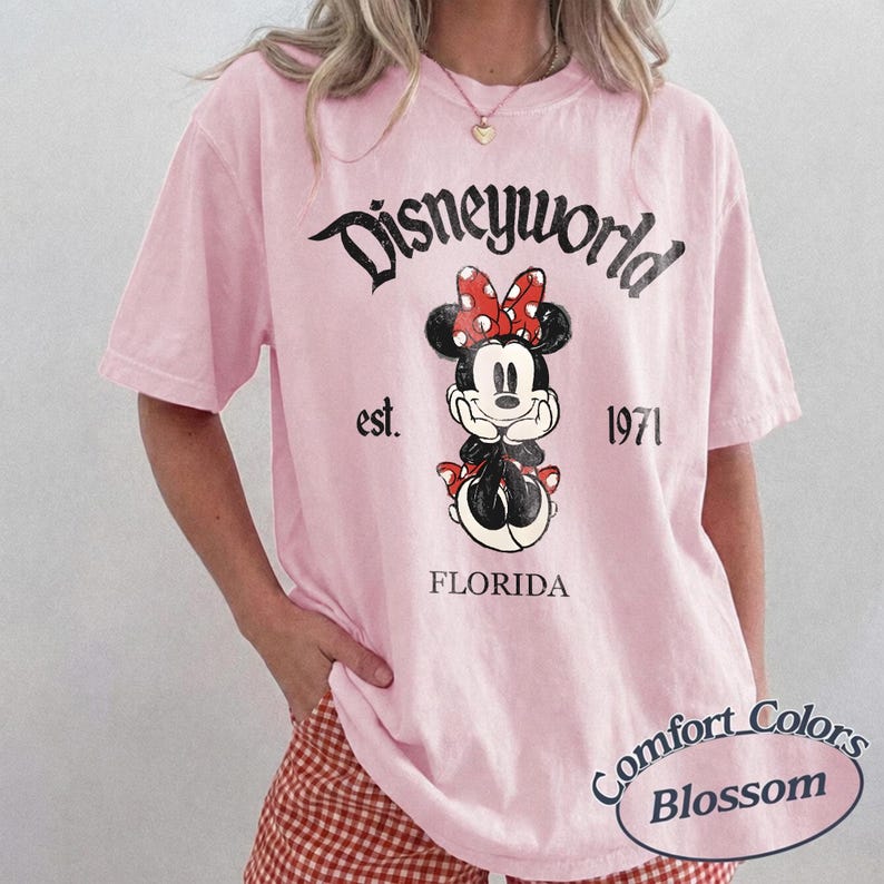 May include: A light pink t-shirt featuring a Minnie Mouse graphic with a red bow and the text "Disneyworld est. 1971 Florida". The shirt is from Comfort Colors.