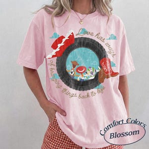 May include: A light pink short-sleeve t-shirt featuring a graphic of a black tire framing cartoon characters. The text reads Love has ways of bringing things back to life. A red cowboy hat and a red cowboy boot are positioned near the tire graphic.