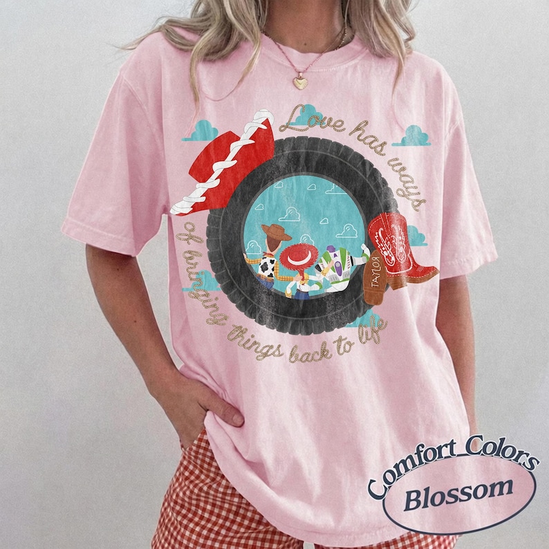 May include: A light pink short-sleeve t-shirt featuring a graphic of a black tire framing cartoon characters. The text reads Love has ways of bringing things back to life. A red cowboy hat and a red cowboy boot are positioned near the tire graphic.