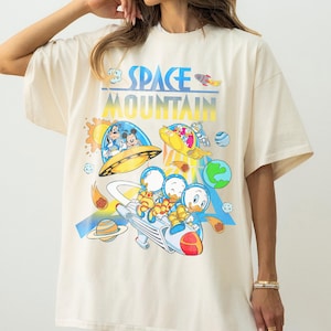 Pu&ograve; includere: T-shirt oversize color crema con una grafica colorata di personaggi dei cartoni animati in un design a tema spaziale. Il testo "SPACE MOUNTAIN" &egrave; stampato in alto in blu e giallo. La maglietta ha maniche corte e una vestibilit&agrave; ampia.