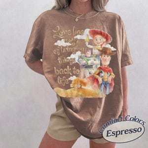 K&ouml;nnte beinhalten: Ein braunes Kurzarm-T-Shirt mit einem Grafikdruck der Toy Story-Figuren Woody, Buzz Lightyear und Jessie. Der Text lautet Love has ways of bringing things back to life. Das T-Shirt hat einen Rundhalsausschnitt und eine lockere, weite Passform.