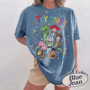 May include: Blue jean-colored t-shirt featuring a colorful Toy Story graphic. The design includes characters like Woody, Buzz Lightyear, and Rex, with the words "Toy Story" in rainbow colors. The shirt is labeled "Comfort Colors Blue Jean".