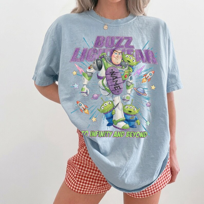 May include: A light blue t-shirt featuring a graphic of Buzz Lightyear and three green aliens. The text reads BUZZ LIGHTYEAR and TO INFINITY AND BEYOND, with a purple oval containing the word NAME. The design includes rockets, stars, and planets.