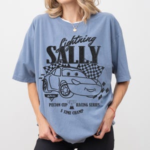 May include: A pale blue t-shirt with a graphic of the character Sally from the film Cars. The design includes the text "Lightning Sally", a chequered flag, and "Piston Cup Racing Series".