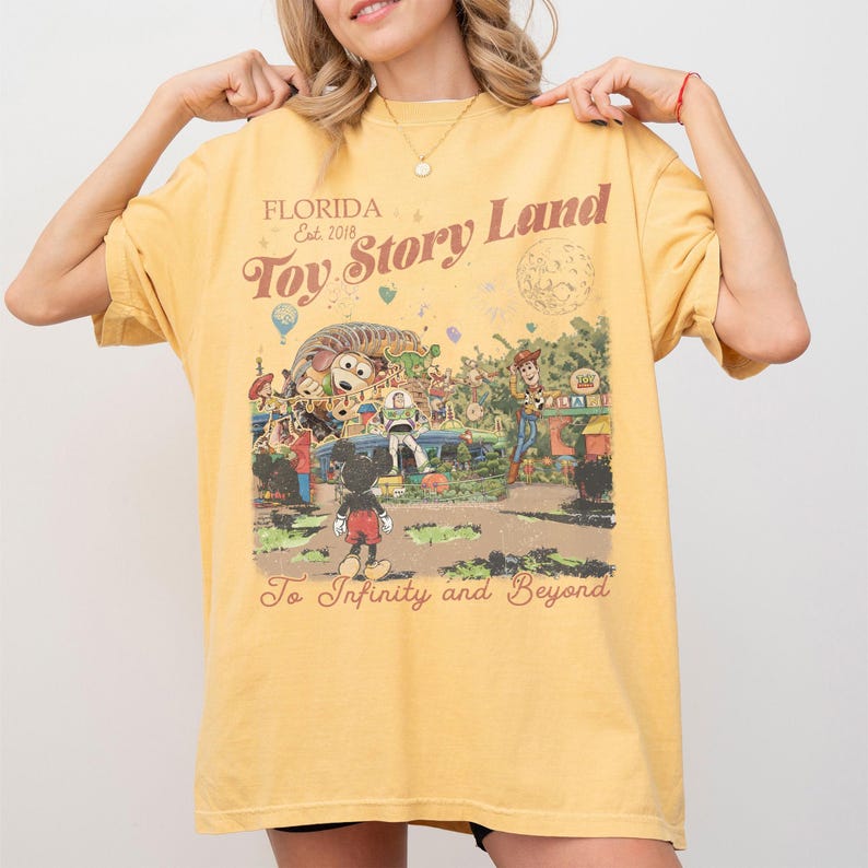 Peut inclure: T-shirt jaune moutarde avec un motif de Toy Story Land. Le design comprend des personnages du film, le texte "Toy Story Land" et "To Infinity and Beyond". Le t-shirt est de coupe ample.
