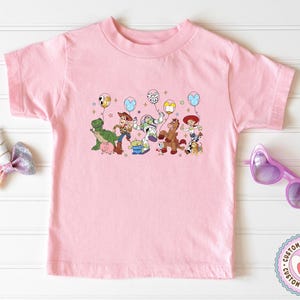 May include: A light pink t-shirt featuring a colorful graphic of characters from the Toy Story movie, including Woody, Buzz Lightyear, and Rex. The characters are holding balloons. A unicorn horn hair bow and heart-shaped sunglasses are also in the image.