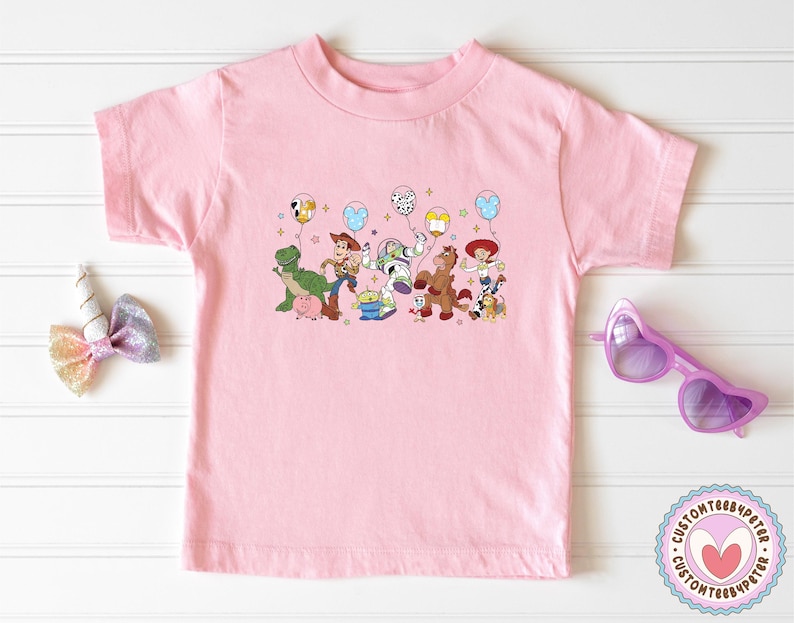 May include: A light pink t-shirt featuring a colorful graphic of characters from the Toy Story movie, including Woody, Buzz Lightyear, and Rex. The characters are holding balloons. A unicorn horn hair bow and heart-shaped sunglasses are also in the image.