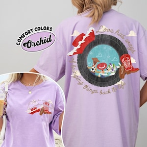 May include: A light purple t-shirt featuring a graphic on the back and front. The back graphic shows a tyre framing cartoon characters, a red hat, and a red cowboy boot with the text Love has ways of bringing things back to life.