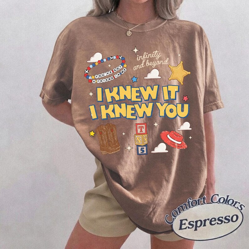 May include: An espresso-colored t-shirt featuring a graphic print with the text "I KNEW IT I KNEW YOU", "infinity and beyond", and "YOU'VE GOT A FRIEND IN ME". The design includes illustrations of a beaded bracelet, a star, cowboy boots, blocks, and a hat.