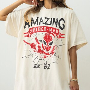 Pode incluir: Camiseta creme com um gr&aacute;fico desgastado do Homem-Aranha. O design inclui as palavras "AMAZING SPIDER-MAN" em preto e vermelho, com "Est. '62" abaixo da imagem do Homem-Aranha.