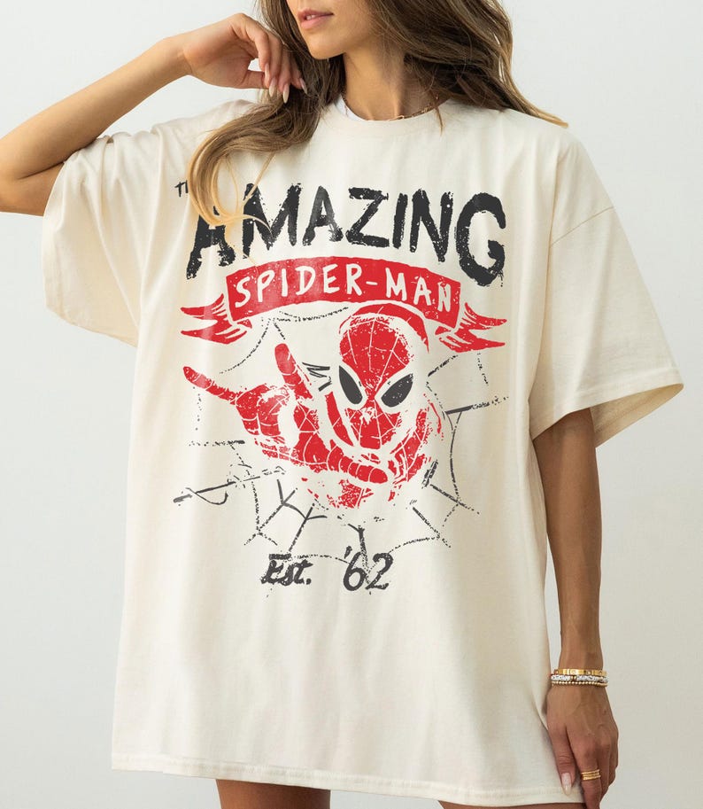 Pode incluir: Camiseta creme com um gr&aacute;fico desgastado do Homem-Aranha. O design inclui as palavras "AMAZING SPIDER-MAN" em preto e vermelho, com "Est. '62" abaixo da imagem do Homem-Aranha.