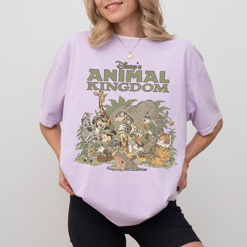 Puede incluir: Camiseta lila claro con el texto "Disney's Animal Kingdom" en verde. El gr&aacute;fico incluye animales de dibujos animados como una cebra, una jirafa, un elefante, un tigre y Mickey Mouse. Estilo casual.