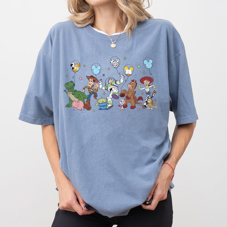 May include: A light blue t-shirt featuring a colorful graphic of animated characters from a popular movie. The design includes characters like a dinosaur, a pig, a cowboy, a space ranger, and a horse, along with balloons.