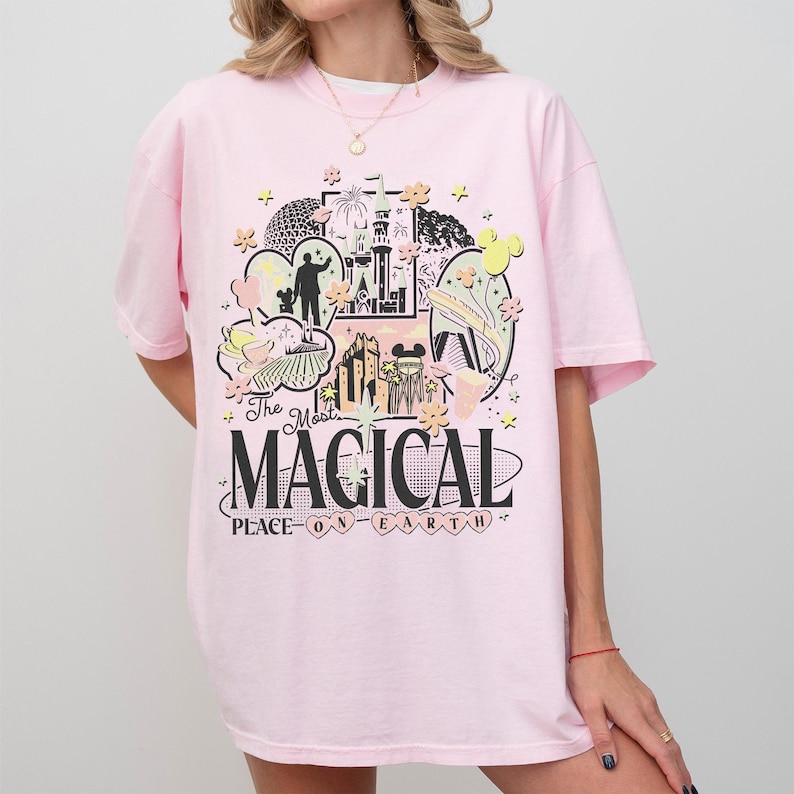 May include: A pale pink t-shirt with a colourful graphic. The design features a castle, a person's silhouette, and the words "The Most MAGICAL PLACE ON EARTH" in a playful font. The t-shirt has short sleeves and a round neck.