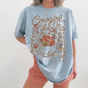 Puede incluir: Una camiseta de manga corta azul claro con un estampado gr&aacute;fico que incluye el texto Cowgirl Like Me y One to Know One. El dise&ntilde;o presenta un borde de cuerda, un sombrero de vaquero rojo, una herradura, un paisaje des&eacute;rtico y notas.