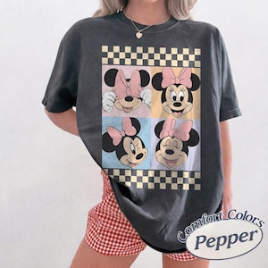 May include: Dark gray t-shirt featuring four panels with Minnie Mouse faces, each with a pink bow. The design includes a checkered border and pastel color blocks. The shirt has the text "Comfort Colors Pepper" on the bottom right.