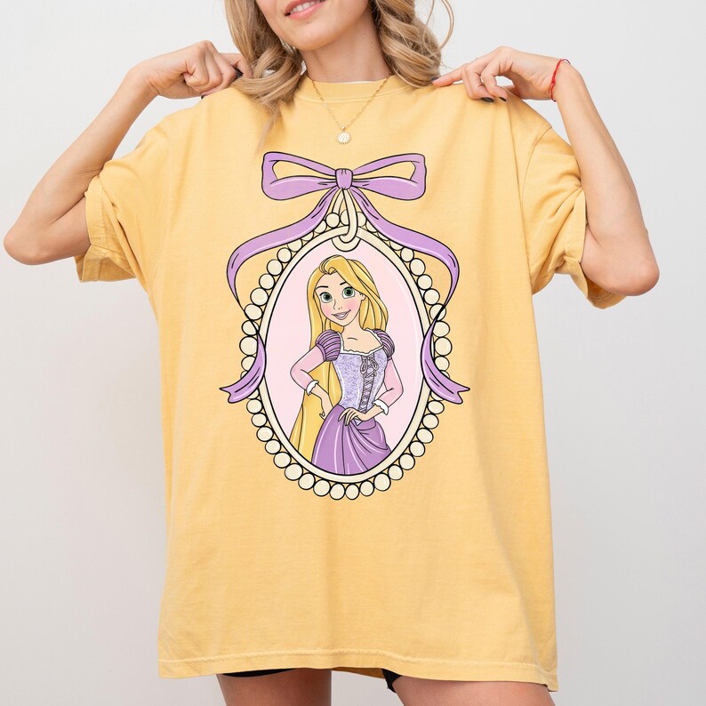 May include: Yellow t-shirt featuring a framed illustration of a cartoon princess with long blonde hair, wearing a purple dress. The frame is oval-shaped, outlined with pearls, and adorned with a purple bow and ribbons.